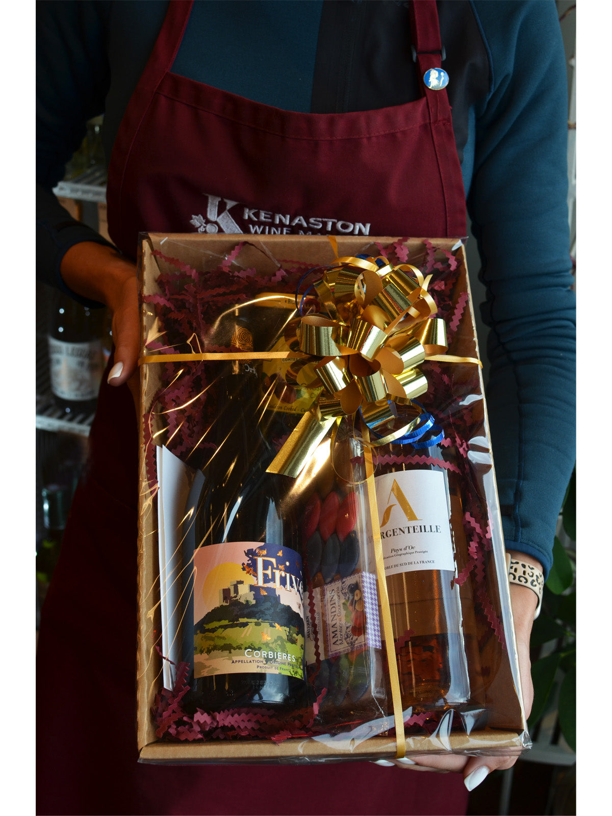 Person holding a wrapped gift box (clear film/gold bow & ribbon) with wine and food, wearing a maroon apron with 'Kenaston Wine Market' branding.