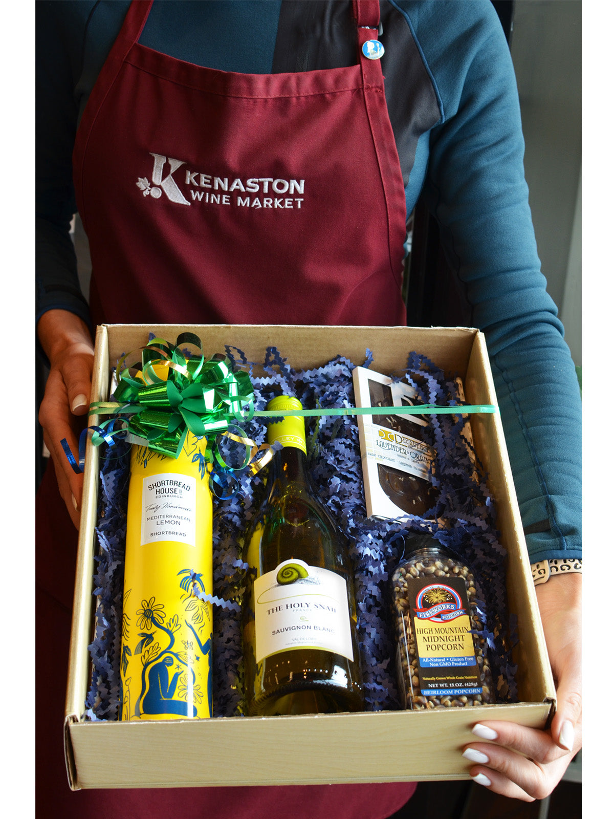 Person wearing a Kenaston Wine Market apron holding a gift box with wine and popcorn kernels.
