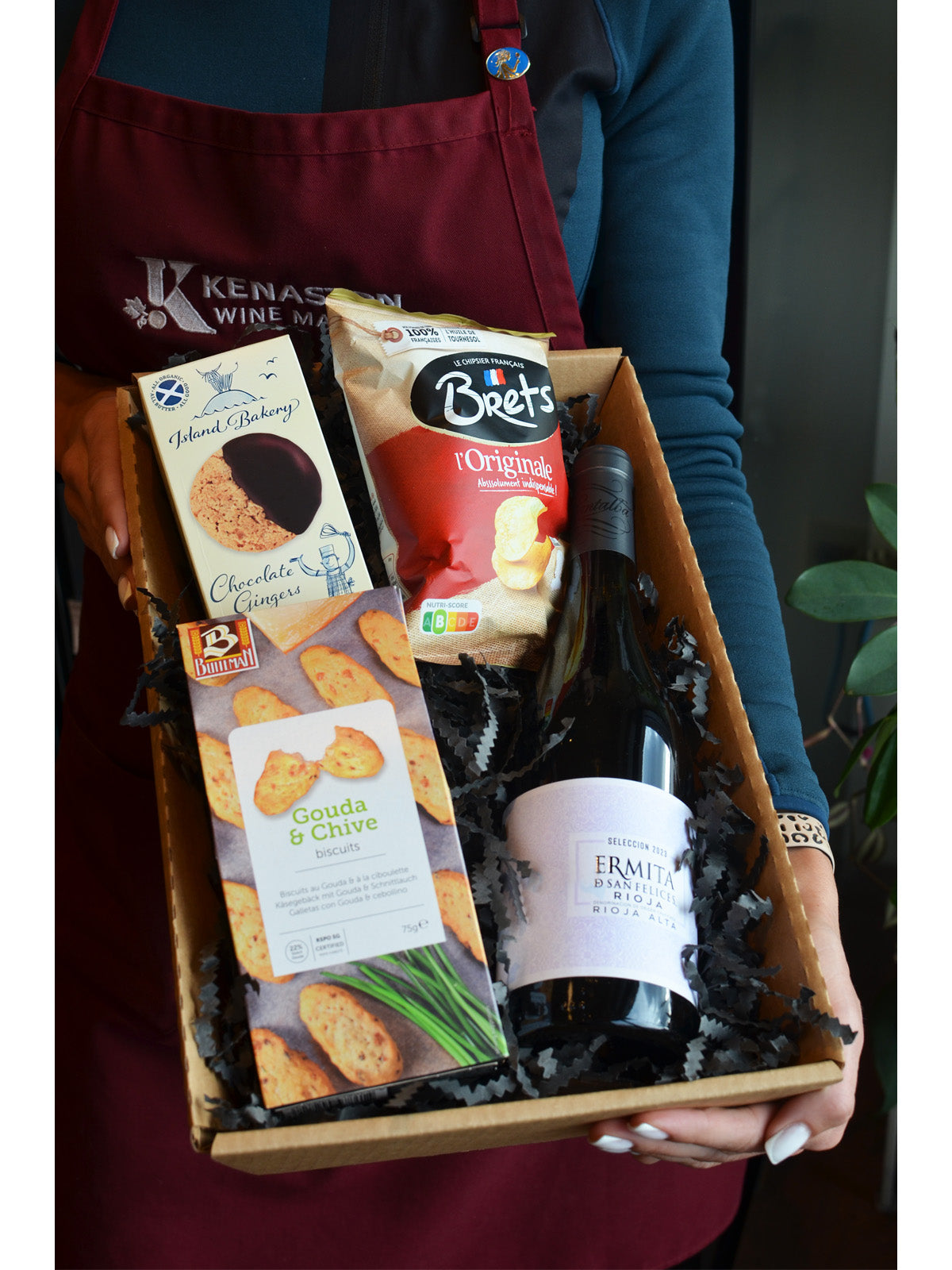 Person holding a gift box with wine, crackers, ginger biscuits and Bretts potato chips.
