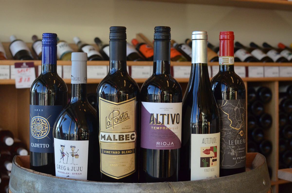 Bottles of Altivo Rioja Tempranillo, Dievole 'Le Due Arbie' Chianti Superiore, La Posta 'Vineyard Blend' Malbec, Complices de Loire 'Altitude' Pinot Noir, Greg and Juju Red, Lungarotti 'Cadetto' Rosso sitting on top of a barrel.