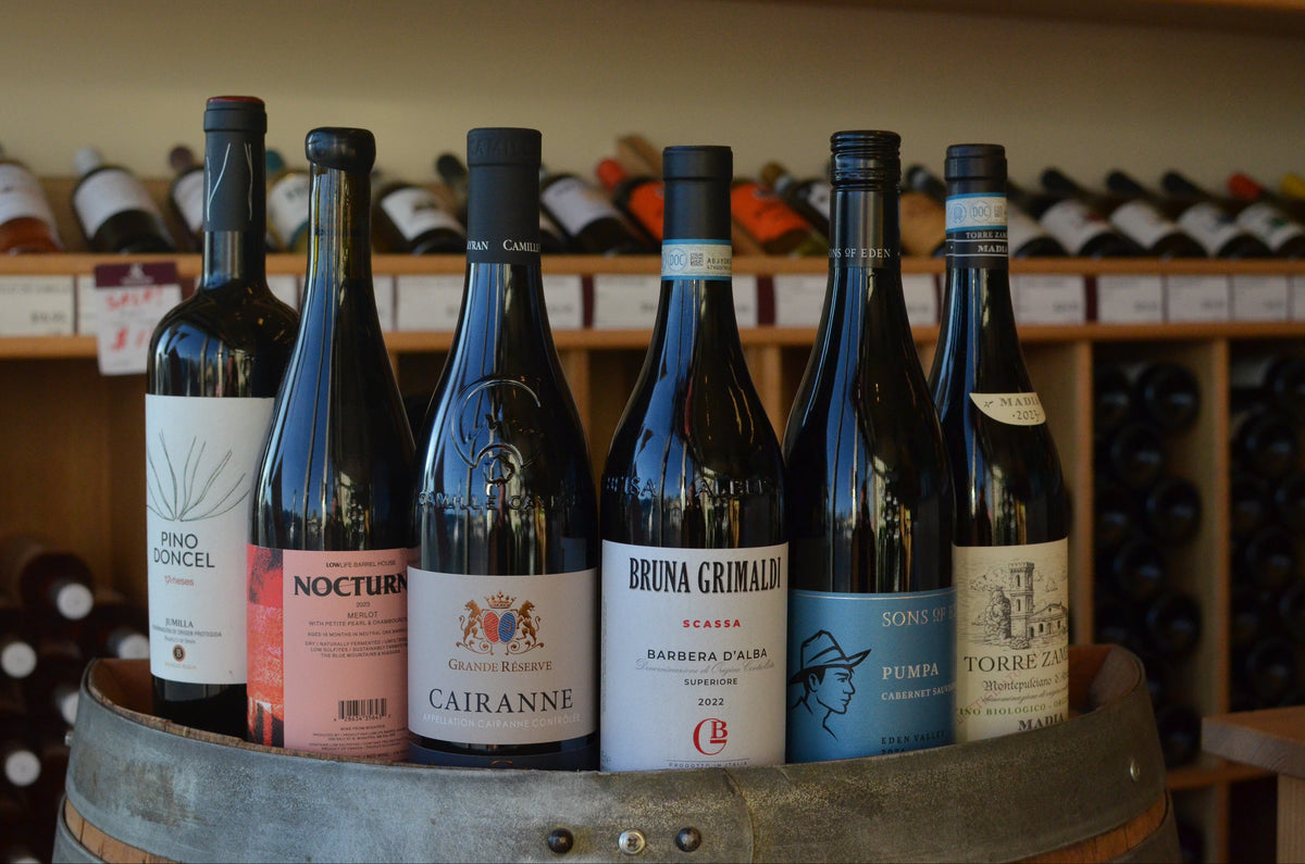 Bottles of Sons of Eden 'Pumpa' Cabernet Sauvignon, Torre Zambra 'Madia' Montepulciano, Low Life "Nocturne" Merlot, Camile Cayran "Le Chene Noir", Bodgeas Bleda "12 Meses" Monastrell Syrah, Bruna Grimaldi "Scassa" Barbera d'Alba sitting on top of a barrel. 