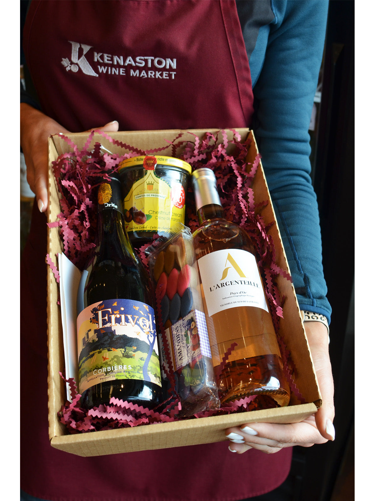 Person holding a gift box with wine and food, wearing a maroon apron with 'Kenaston Wine Market' branding.